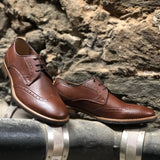 A tan vegan leather brogue dress shoe, lace up with 3 eyelets. Tan and dark brown sole with rubber grip for traction. Shown against a cement background.