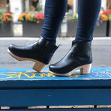 A pair of black vegan leather clog booties on a woman's feet. Ankle height shaft with pull tab in back. Inside zipper closure. Blonde wooden sole. Staples around outsole to connect material to sole. Street in background.